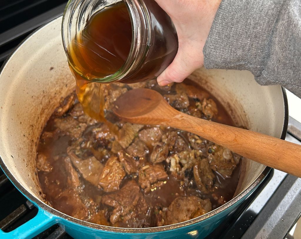 Enameled cast iron pot with mason jar of beef stock being added to meat and onion mixture.