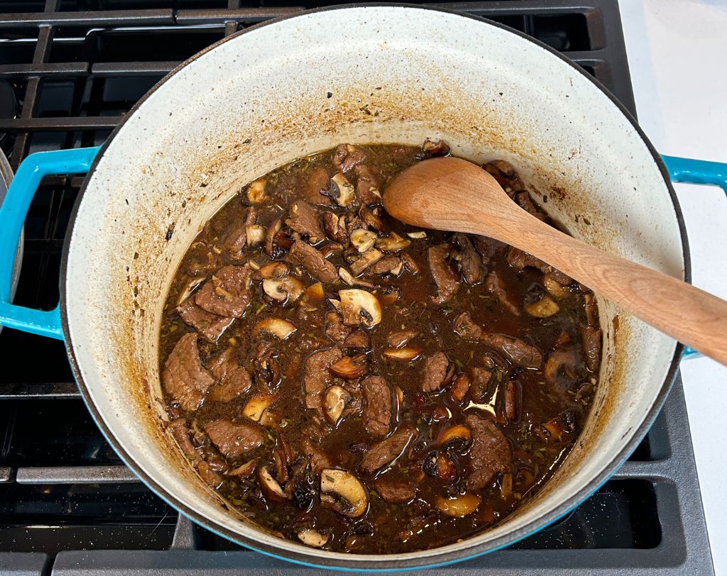 Enameled cast iron pot on stove top with wooden spoon, meat, onion, herb and garlic mixture reduced and tender.