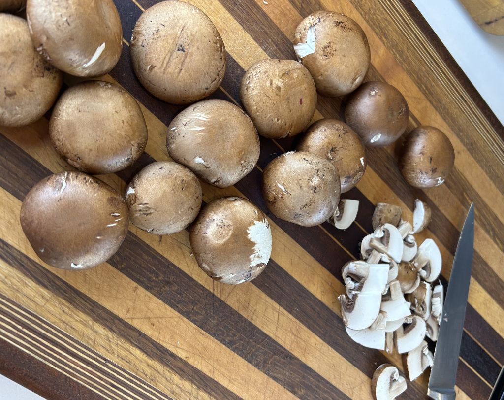 Wood grain cutting board with utility knife, whole and sliced cremini mushrooms.