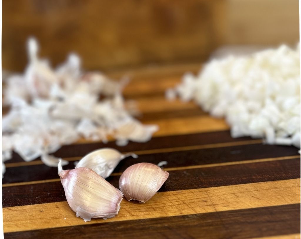 Wood cutting board with three cloves of garlic and chopped onions blurred in the background.