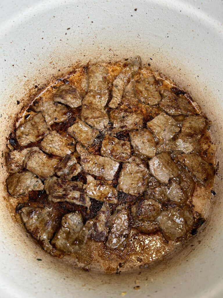 Enameled cast iron pot with golden brown beef strips cooking inside.