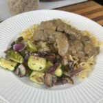 White plate with buttered noodles, Delicious Beef Stroganoff and roasted vegetables with dried pasta in the background.