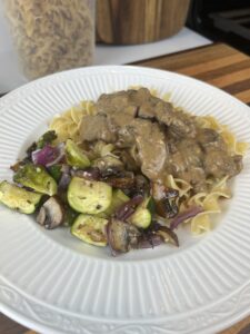 White plate with buttered noodles, Delicious Beef Stroganoff and roasted vegetables with dried pasta in the background.