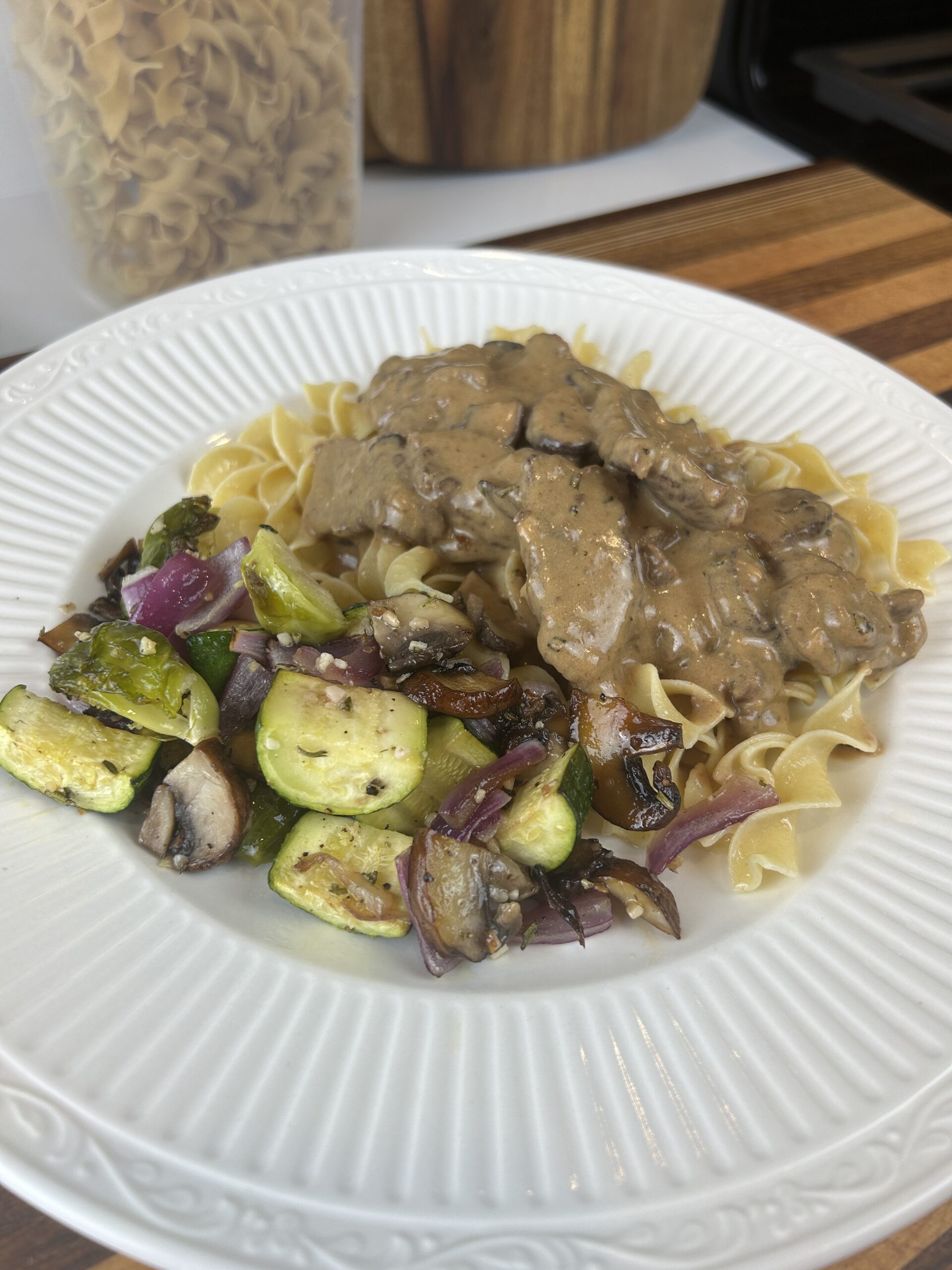 White plate with buttered noodles, Delicious Beef Stroganoff and roasted vegetables with dried pasta in the background.