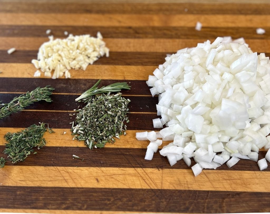 Wooden cutting board with chopped onions, garlic, rosemary and thyme.