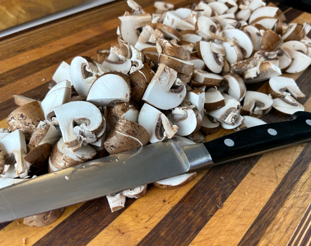 Wood grain cutting board with utility knife and sliced cremini mushrooms.