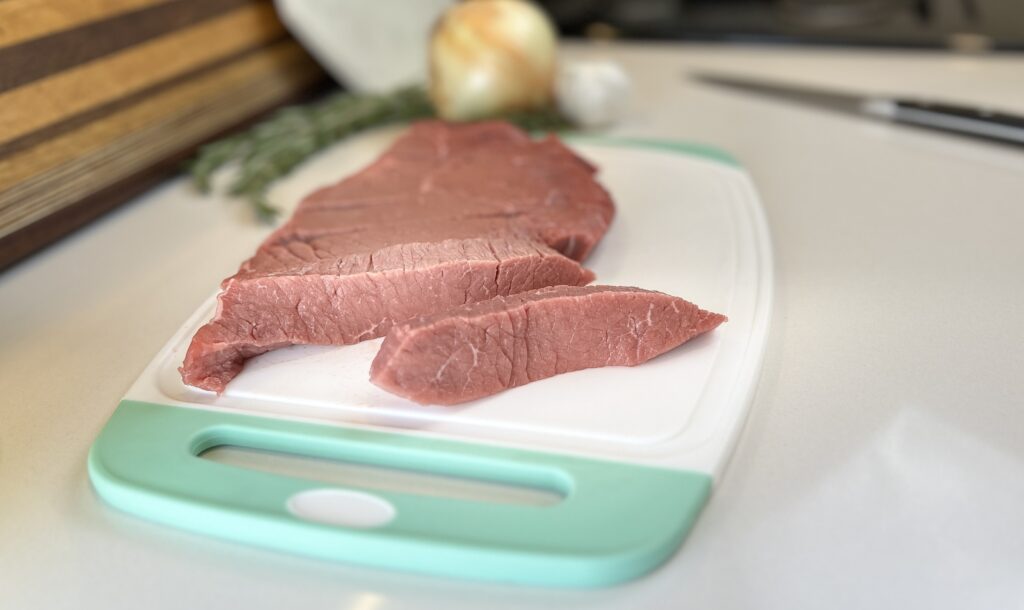 White and aqua cutting board with London broil sliced into large strips.