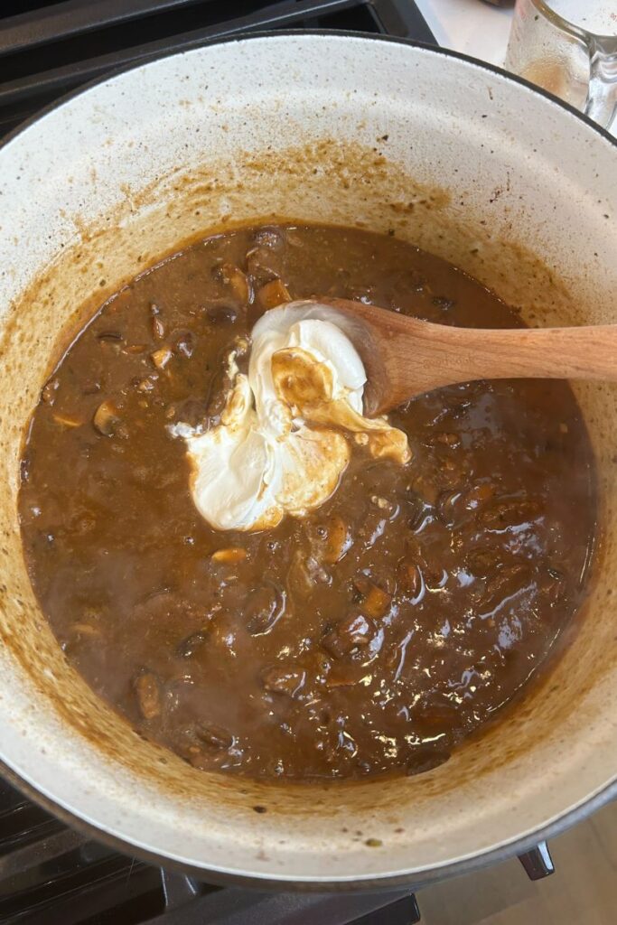 Delicious Beef Stroganoff in enameled cast iron pot with wooden spoon mixing in 3/4 cup of sour cream.