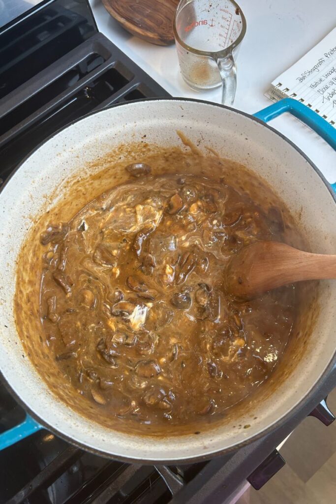 Delicious Beef Stroganoff in enameled cast iron pot with wooden spoon mixing in sour cream.