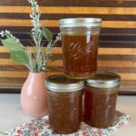 Three Jars of homemade chicken stock in front of a striped wood cutting board with flowers in a coral vace.