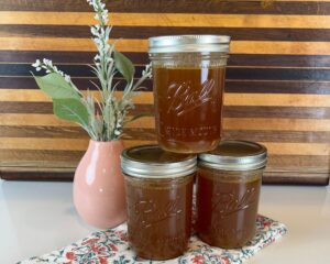 Three Jars of homemade chicken stock in front of a striped wood cutting board with flowers in a coral vace.