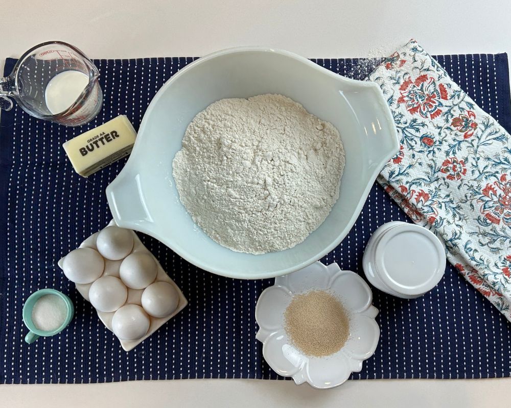 Mixing bowl with flour, white dish with active yeast, white salt container, small dish of sugar, white tray with six eggs all sitting on a navy towel with a coral and navy floral towel on the right. 