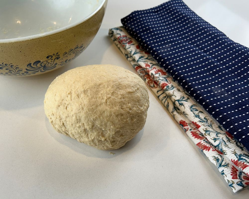 Rough ball of dough on white counter with brown Pyrex bowl and navy tea towel in the background.