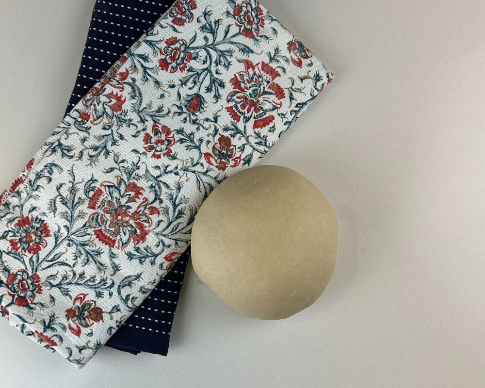 Homemade Challah  dough ball smooth and elastic on white counter with one navy tea towel and one floral tea towel.