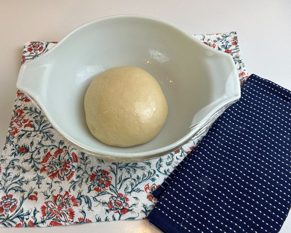 Homemade Challah Dough ball in lightly oiled bowl with tea towel in background.