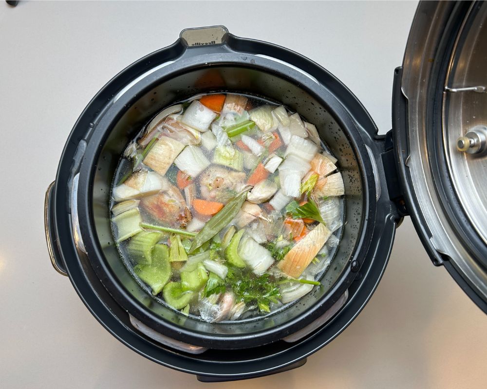 Pressure cooker pot with Homemade Chicken Stock ingredients. Roasted chicken wings, carrots, celery, onion, garlic and parsley covered in filtered water.