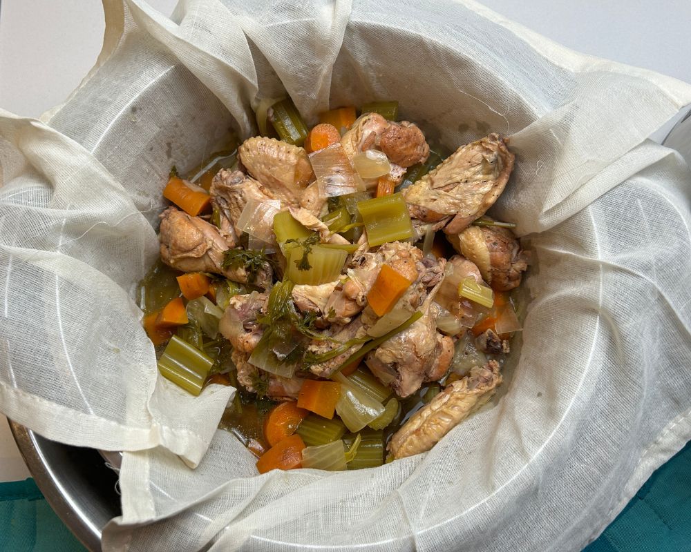 Homemade chicken stock ingredients draining through a cheesecloth lined colander.