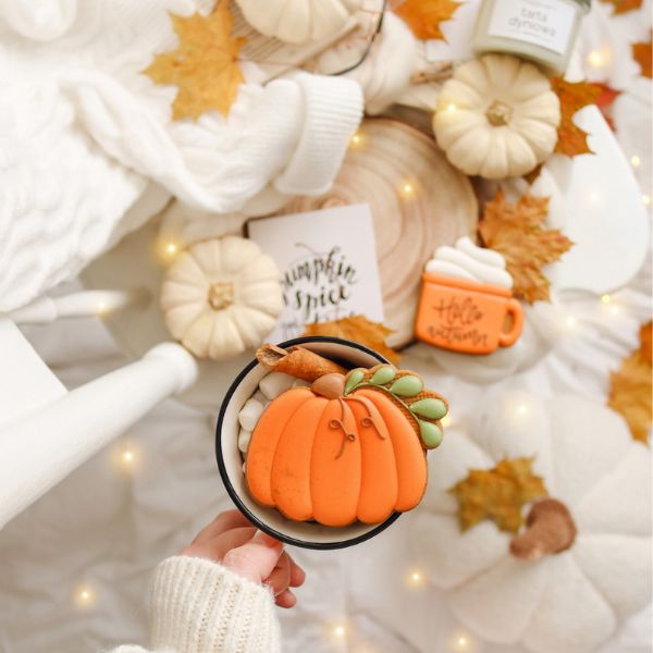 A warm, inviting fall dining setup. Two lit beeswax candles stand in golden holders on a neutral-colored tablecloth. A bouquet of dried hydrangeas in a glass vase sits between large orange and white pumpkins. The table is set with fine china, featuring intricate designs and gold accents, ready for a cozy autumn gathering.