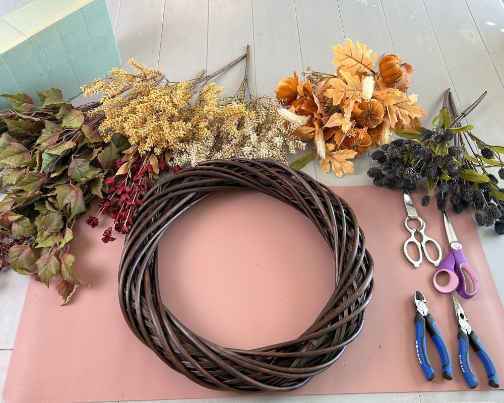 Dark brown vine wreath with various floral pics in yellow, orange, cream, burgundy and blue. Scissors and pliers on mauve work surface.