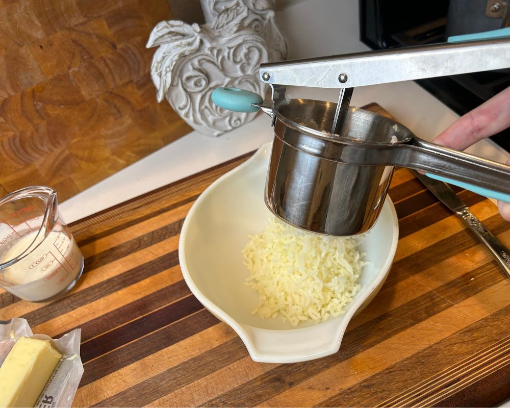 Cooked potatoes in potato ricer basket over serving bowl being pressed through.