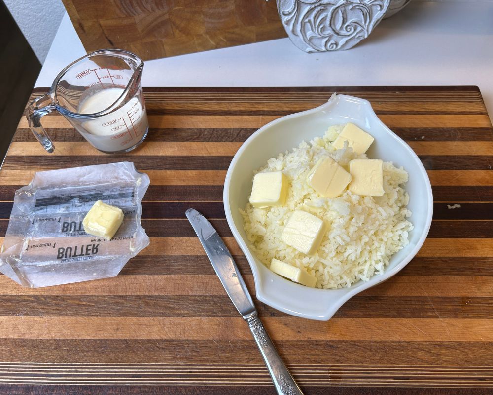 Striped cutting board with dish of mashed potatoes, butter sliced on top and heavy cream in a measuring cup. 