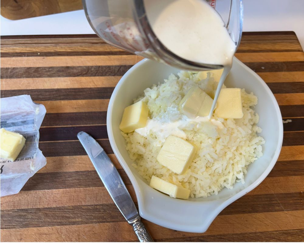 Striped cutting board with dish of mashed potatoes, butter sliced on top and heavy cream being poured into the dish.