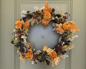 Vine wreath with a mix of floral picks in orange, cream, yellow, burgundy, green and blue. Hanging on sage green door.