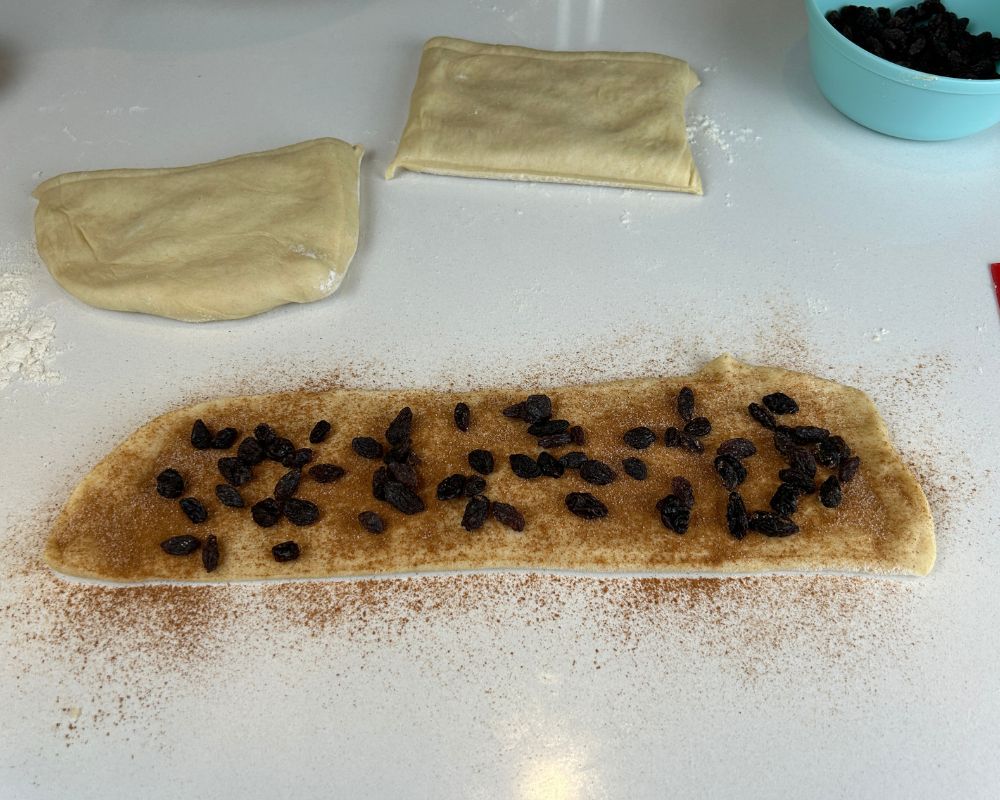 Flat length of Homemade Challah dough covered in cinnamon sugar and raisins.