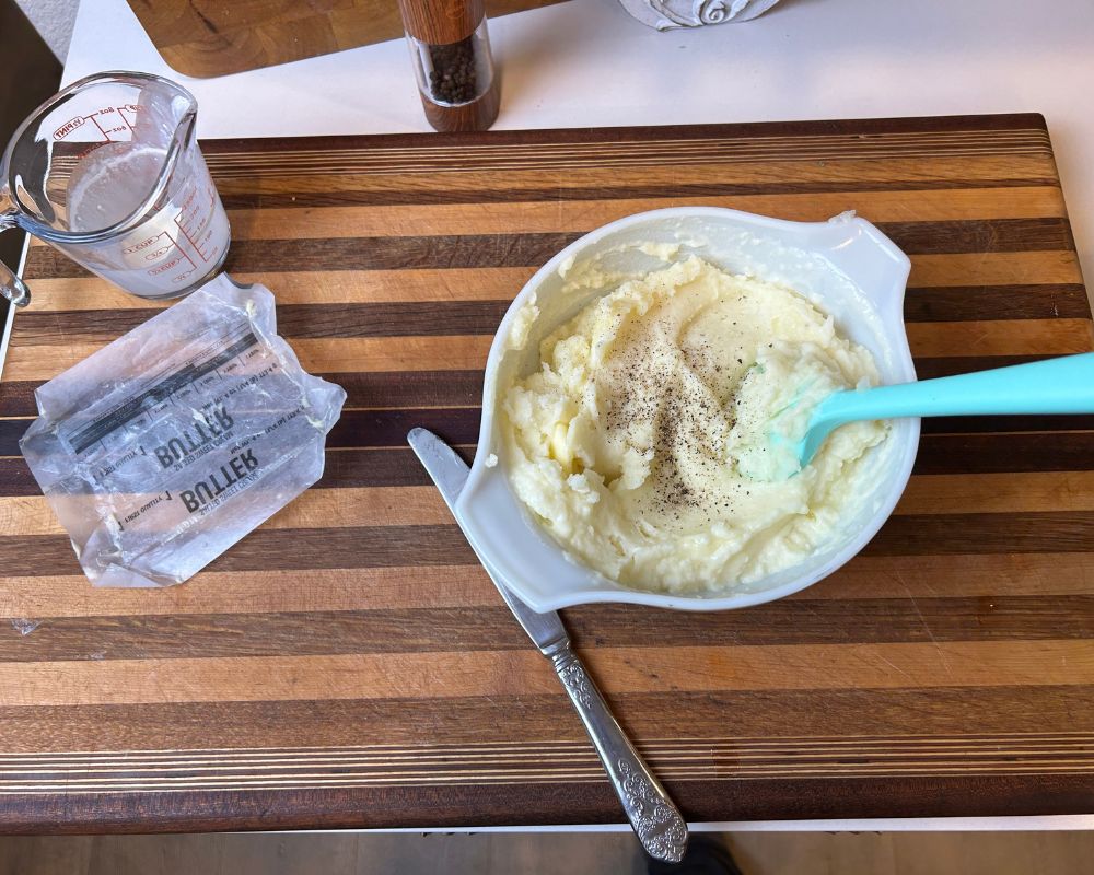 Striped cutting board with dish of mashed potatoes mixed with fresh ground pepper on top.