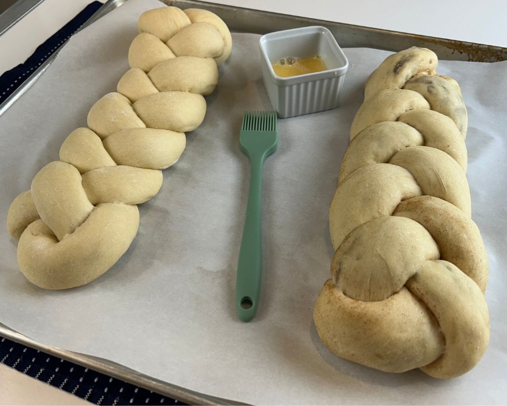 Two Homemade Challah braids with egg wash in a dish and a green silicone brush on a parchment lined baking sheet. 