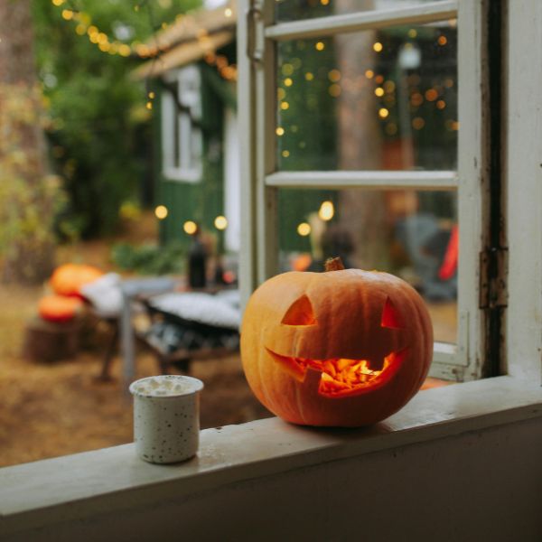 A carved jack-o'-lantern with a glowing candle inside sits on a windowsill, giving off a warm, inviting light. Next to it, there's a small speckled mug. Through the open window, an outdoor scene is visible with soft string lights and cozy seating, featuring a bench with blankets and pillows, creating a charming autumnal evening atmosphere. The outdoor area is surrounded by greenery and trees, adding to the cozy, rustic vibe.