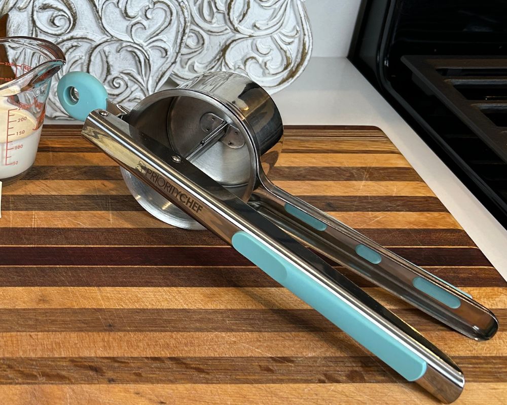 Striped cutting board with carved wooden fruit behind it. A silver and aqua potato ricer is sitting on the cutting board. 