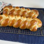 Two Homemade Challah Bread braids on a cooling rack sitting on a blue and white hand towel on a white counter.