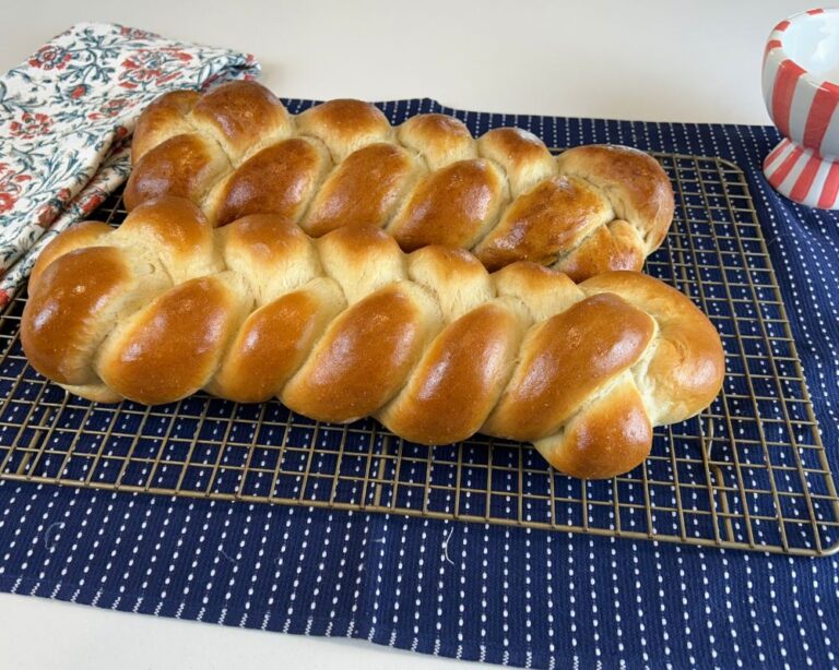 Two Homemade Challah Bread braids on a cooling rack sitting on a blue and white hand towel on a white counter.