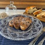 Glass decorative plate on navy blue towel with powder sugar dusted Challah French Toast. Sliced Cinnamon Raisin Challah and butter with toasted pecans in the background.