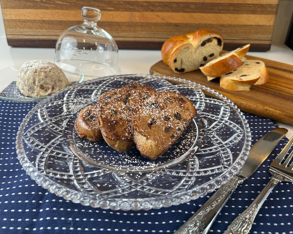 Glass decorative plate on navy blue towel with powder sugar dusted Challah French Toast. Sliced Cinnamon Raisin Challah and butter with toasted pecans in the background.