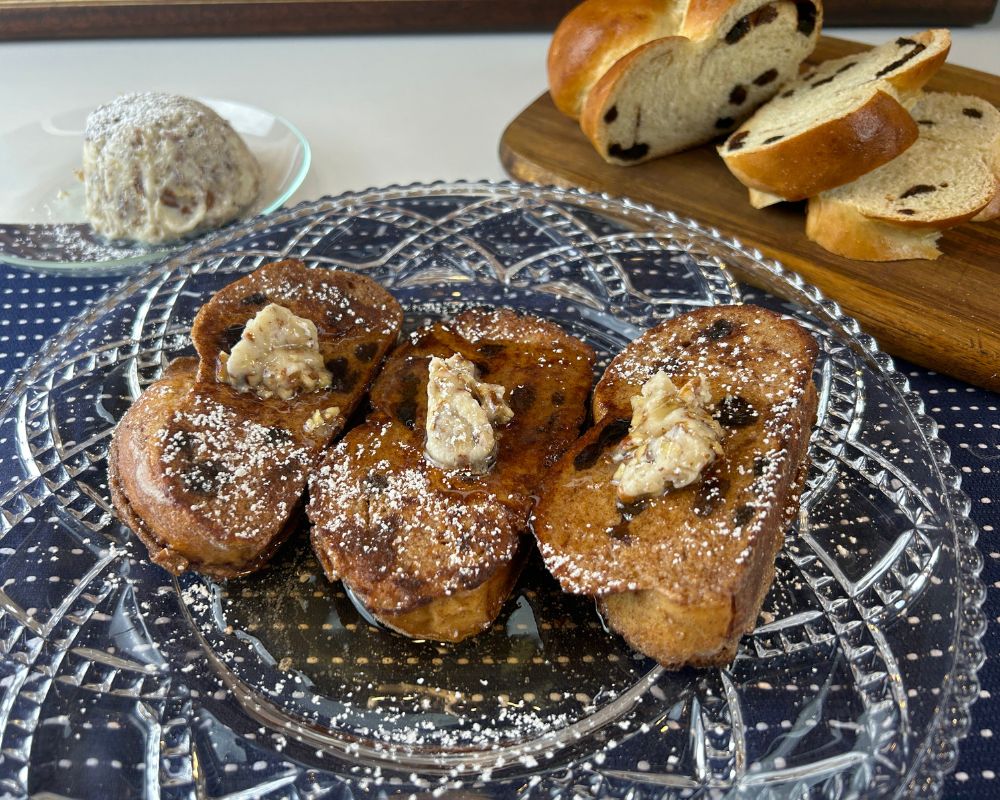 Glass decorative plate on navy blue towel with powder sugar dusted Challah French Toast with butter with toasted pecans on top. Sliced Cinnamon Raisin Challah and butter with toasted pecans in the background.