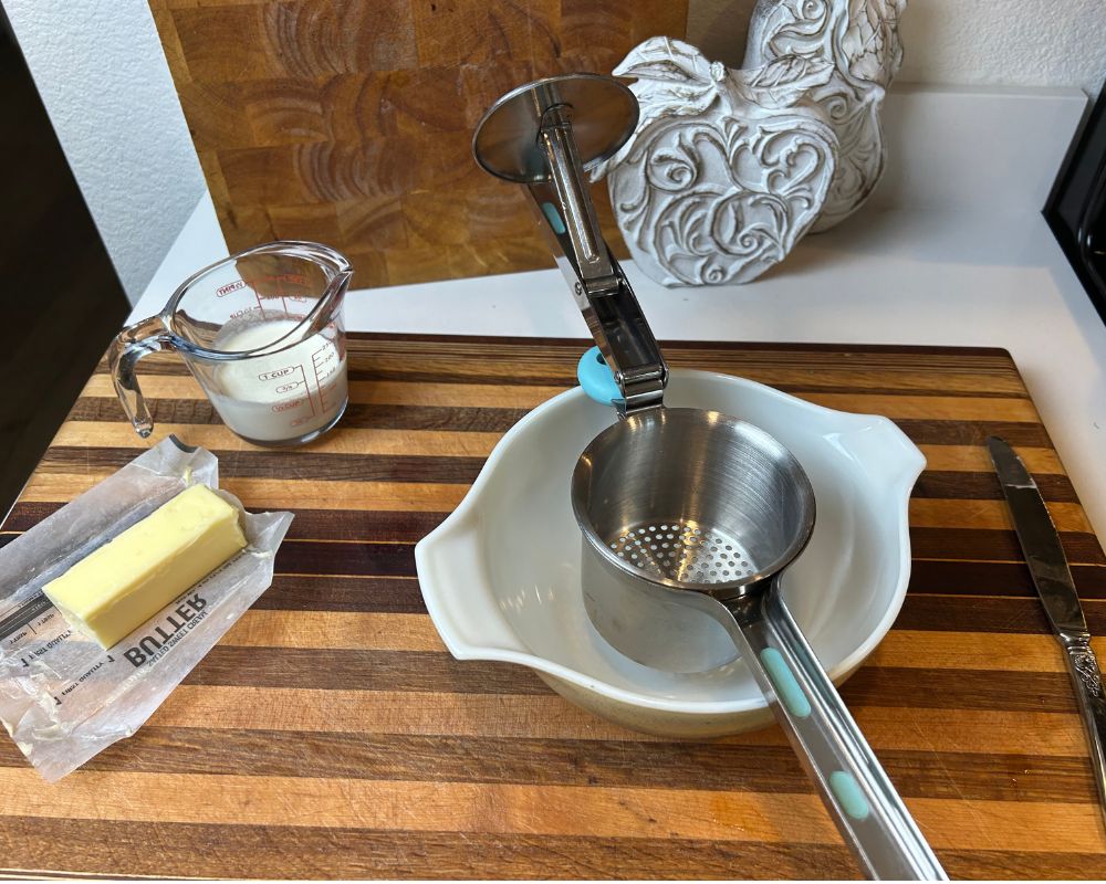 Striped cutting board with butter, heavy cream, serving bowl and potato ricer on top. 