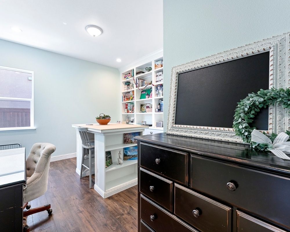 Large black dresser at front entry of craft room with aqua walls, white craft island and wall of built in cabinets and shelves.