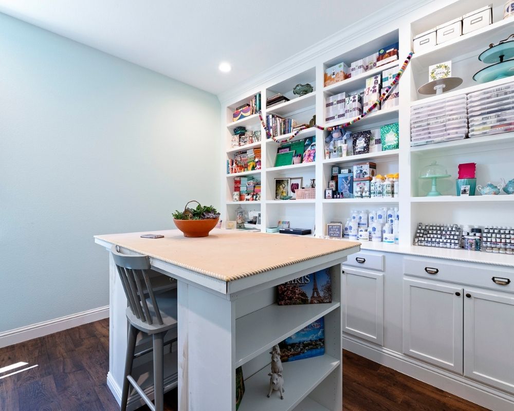 craft room with aqua walls, white craft island and wall of built in cabinets and shelves.