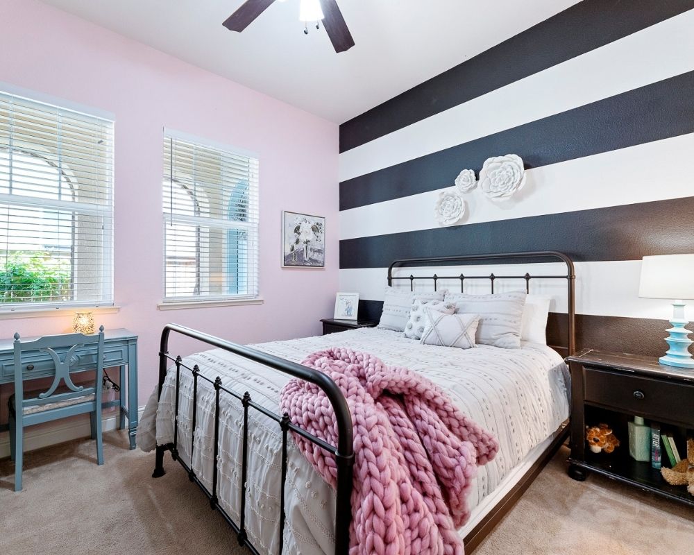 Girls bedroom with pink wall, black and white striped wall with a metal bedframe with light gray bedding. 