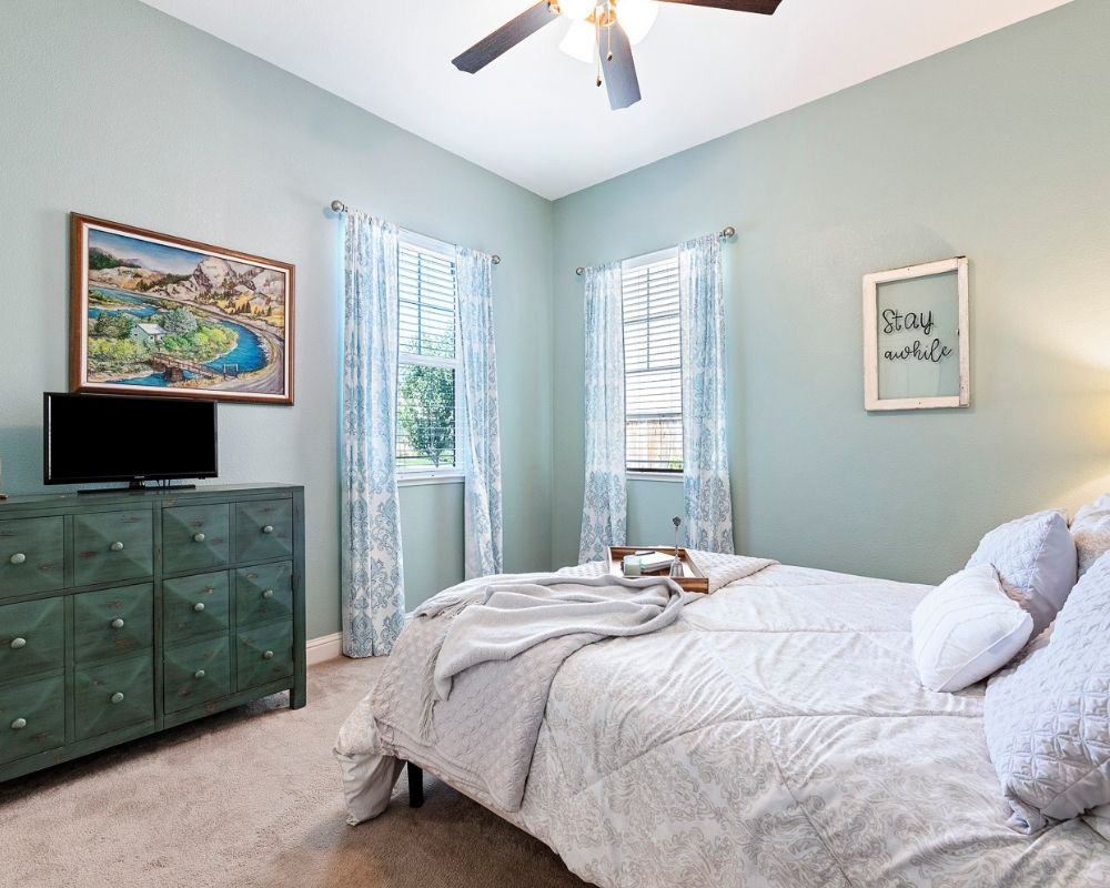 Green walled bedroom with dark green dresser, light gray bedding and windows in the corner. 