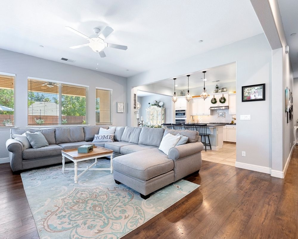 Image of living room with kitchen and dining room in the background. Large gray sectional sofa, blue paisley rug.