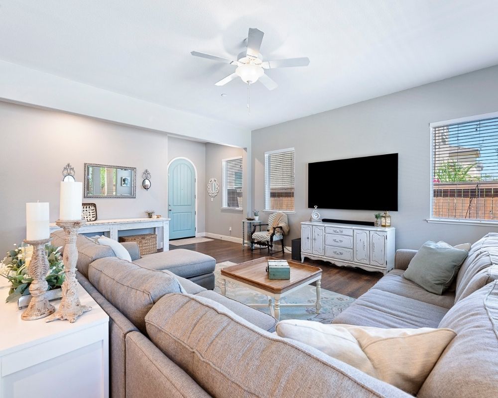 Image of living room and front door with a large gray sectional sofa, blue paisley rug with a tv mounted on the wall over a antique server.