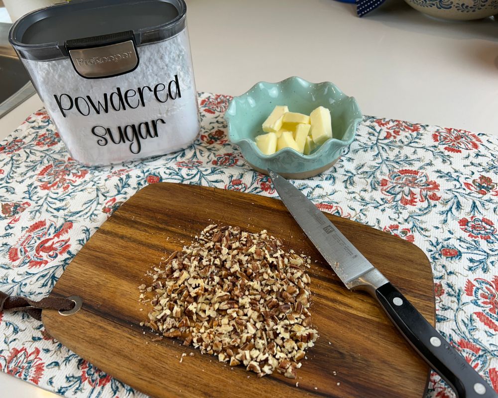 Chopped pecans on a wood cutting board on top of a floral tea towel with a dish of butter and powdered sugar in the background. 