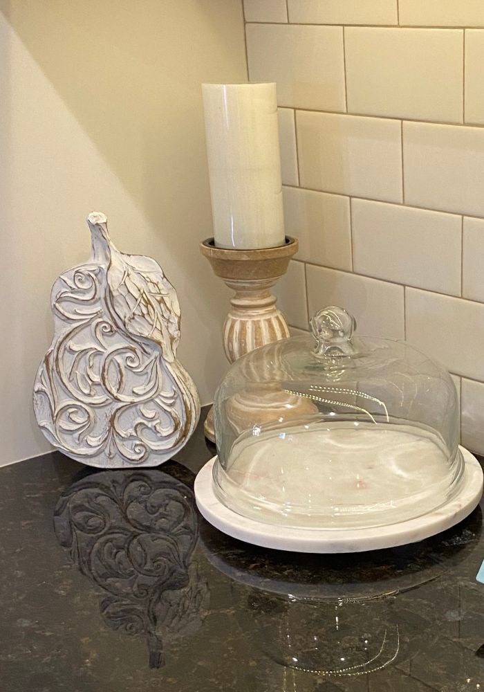 Neutral kitchen vignette with white tile backsplash, glass cake stand, carved decor accent, and soft candlelight on dark countertop
